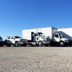 Several vehicles which makes up part of the fleet of Redkey Express trucks, on gravel, shown under a bright blue sky.