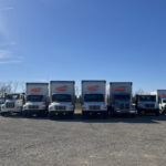 10 vehicles which makes up part of the fleet of Redkey Express trucks, on gravel, shown under a bright blue sky.