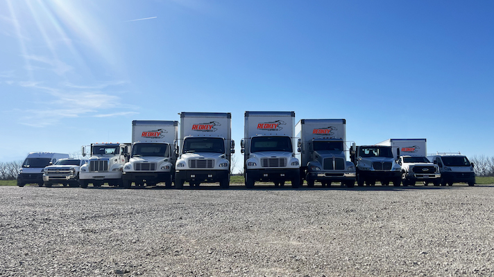 10 vehicles which makes up part of the fleet of Redkey Express trucks, on gravel, shown under a bright blue sky.